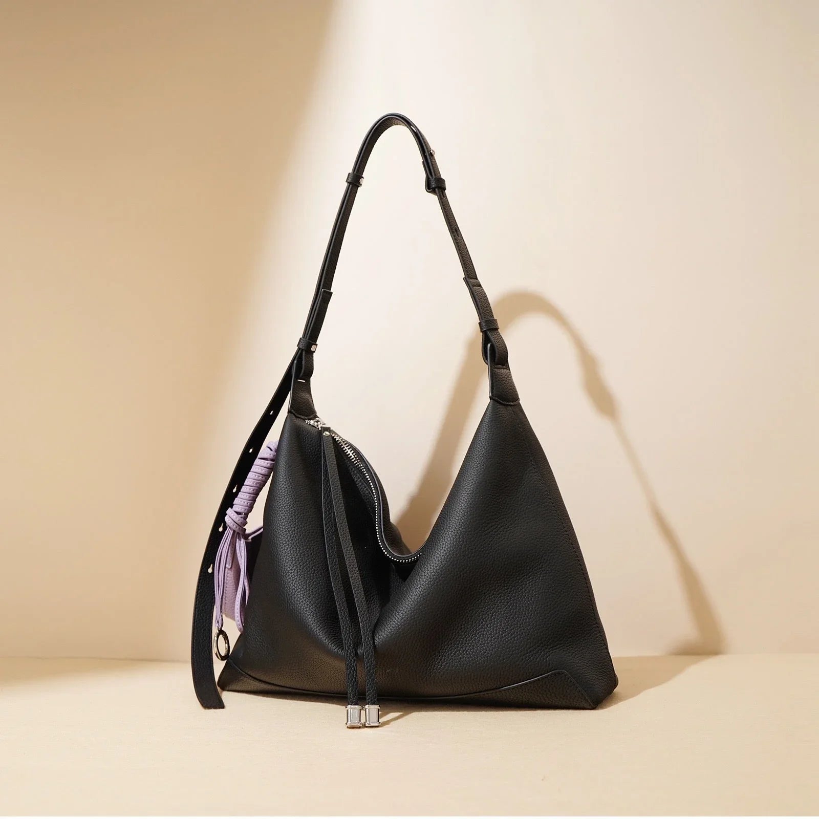 Black leather hobo bag with adjustable strap and lilac accent cord on beige background