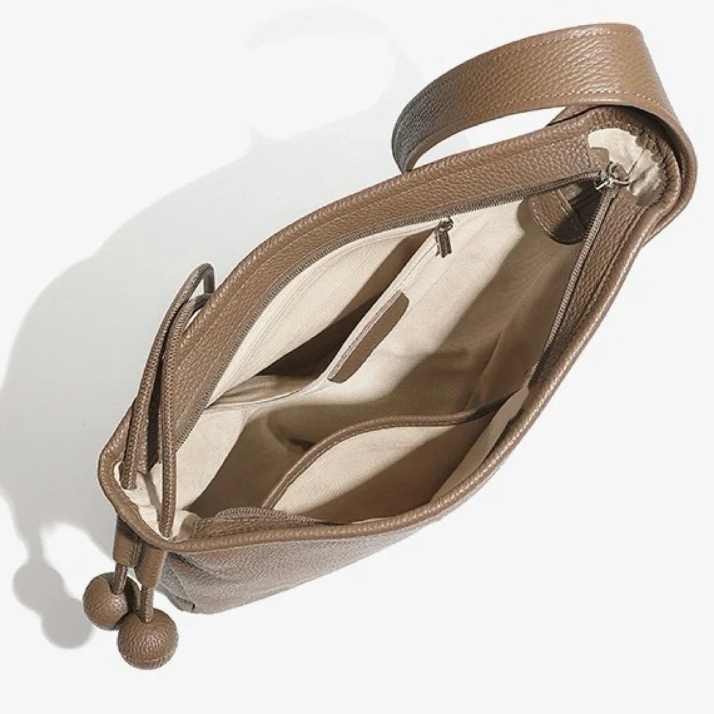 Open brown leather tote bag with beige interior, zip pocket, and double pom pom details