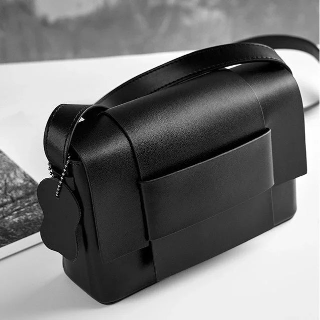 Modern black leather crossbody bag with front pocket, decorative tag on white surface