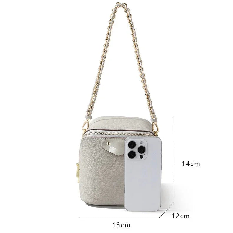 Small beige crossbody bag with chain strap, gold zipper, and phone for size reference, 14x13x12cm.