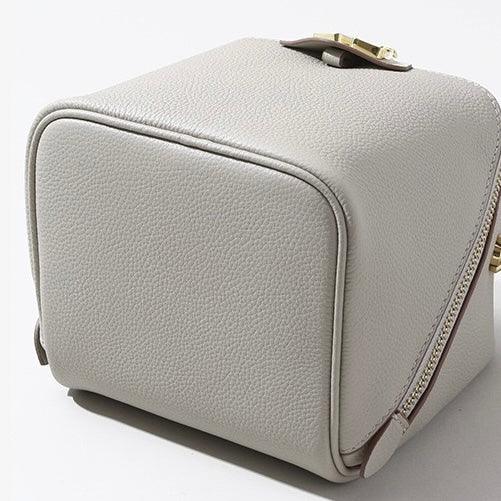 Minimalist light gray leather box purse with zipper and gold clasp on white background