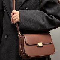 Woman wearing dark coat holding brown leather crossbody bag with gold clasp