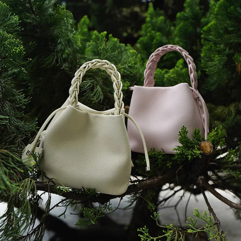 Two leather handbags with braided handles, cream and blush, displayed on greenery outdoors