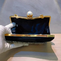 Black evening clutch bag with gold frame, pearl clasp, held open by gloved hand on table
