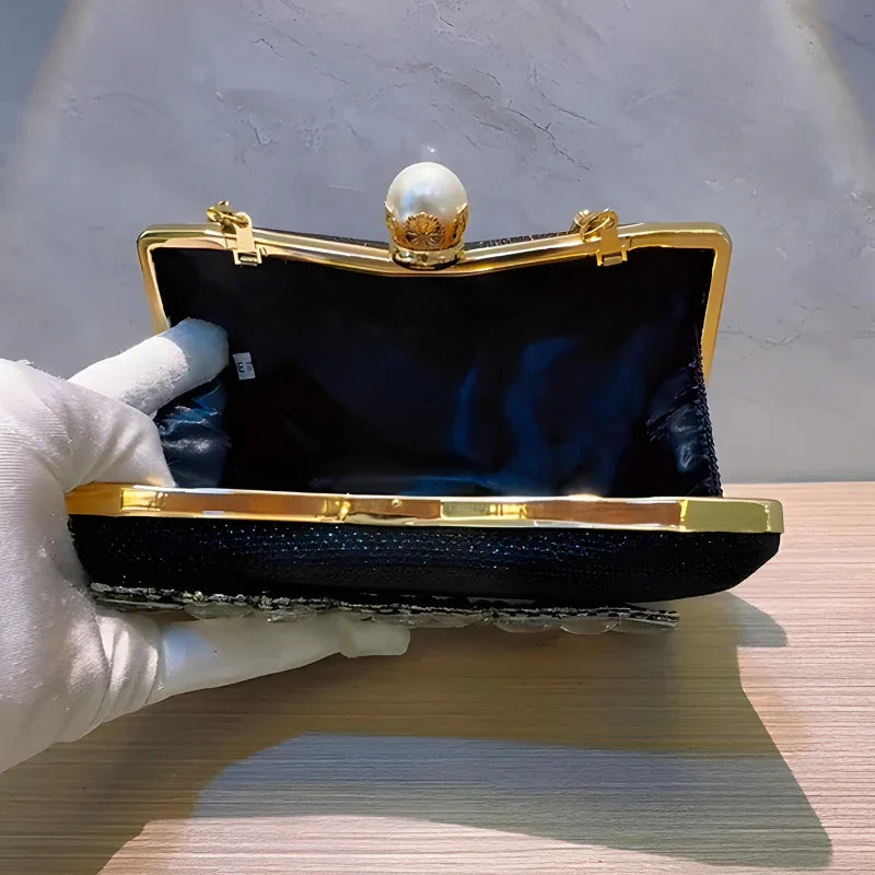 Black evening clutch bag with gold frame, pearl clasp, held open by gloved hand on table