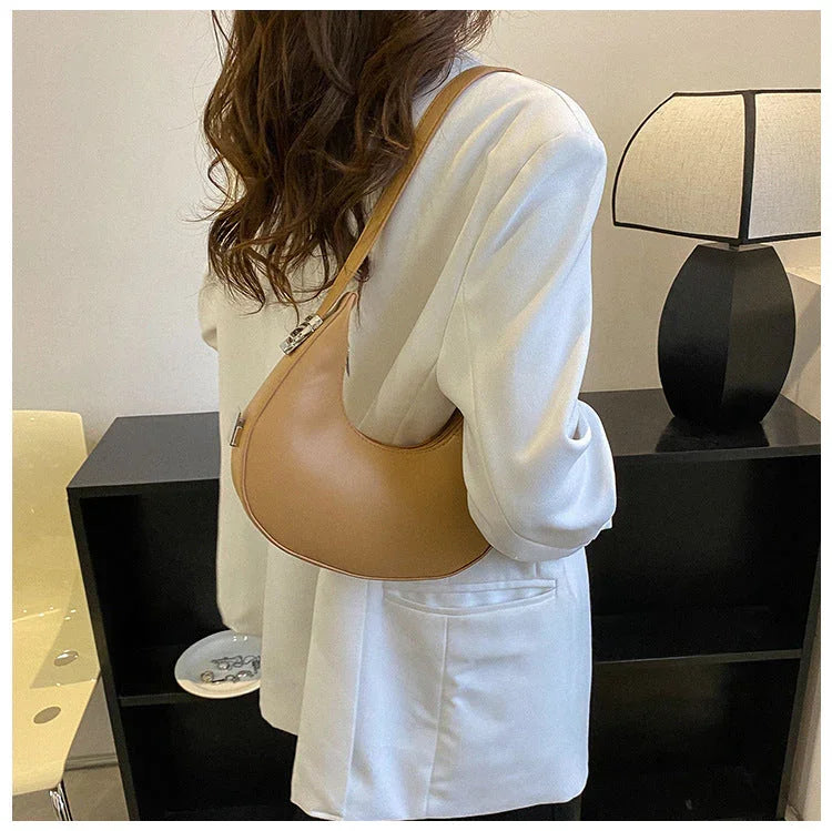 Woman in white blazer carrying a tan crescent shoulder bag indoors by a black lamp