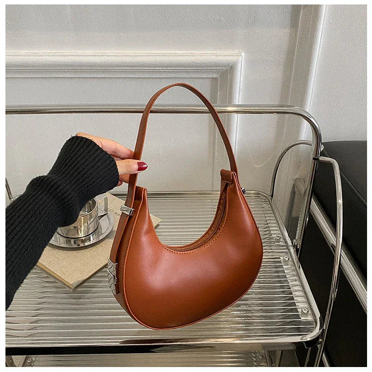 Brown crescent shoulder bag on metal shelf, hand holding handle, minimalist style