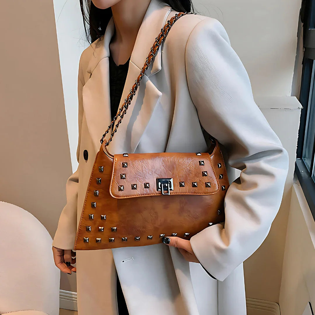 Woman in beige coat holding brown studded leather shoulder bag near window