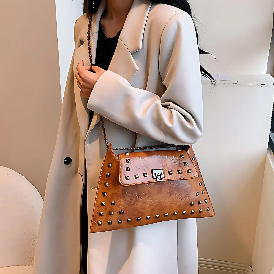 Woman in beige coat holding brown studded leather shoulder bag indoors