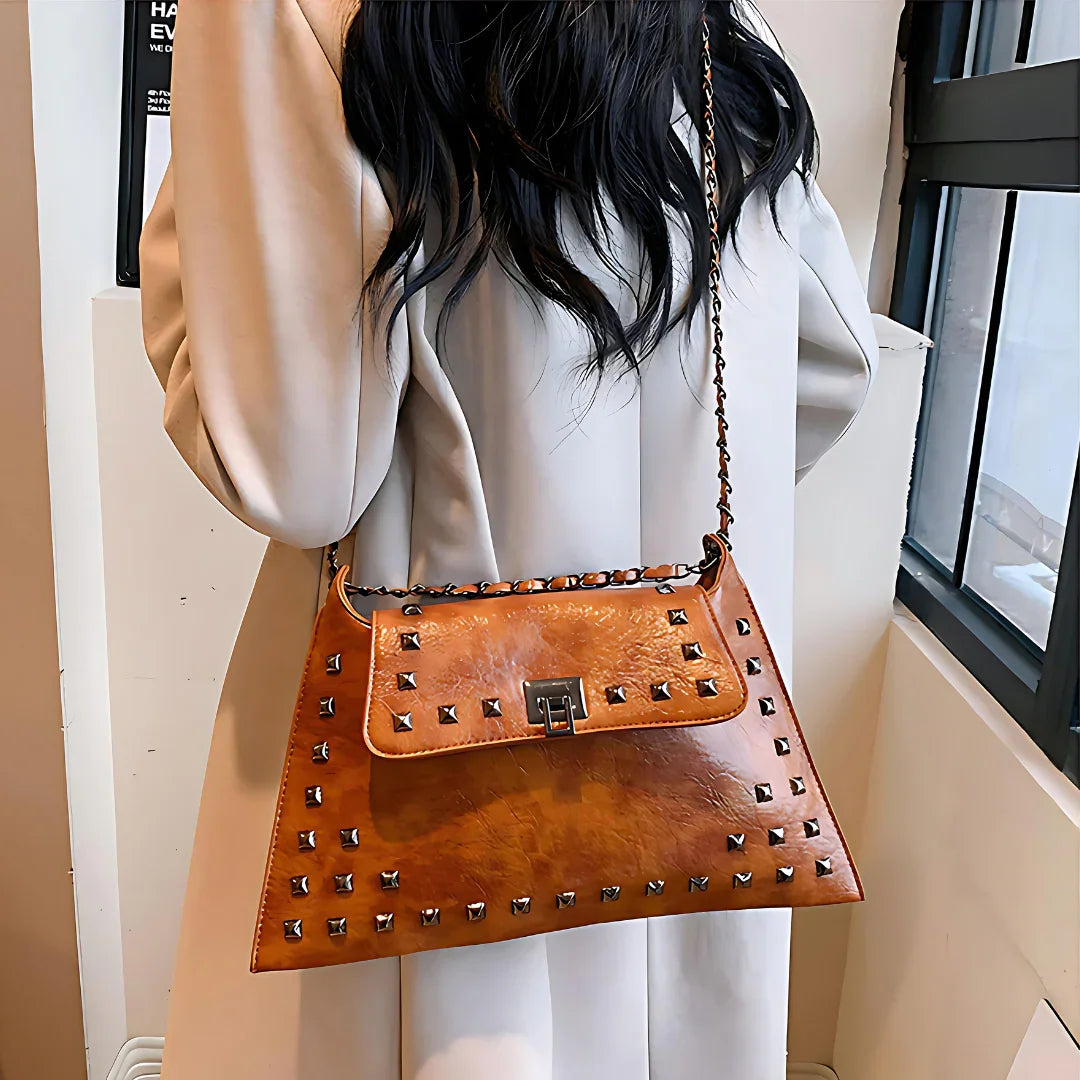 Brown studded leather shoulder bag with chain strap worn over beige coat indoors