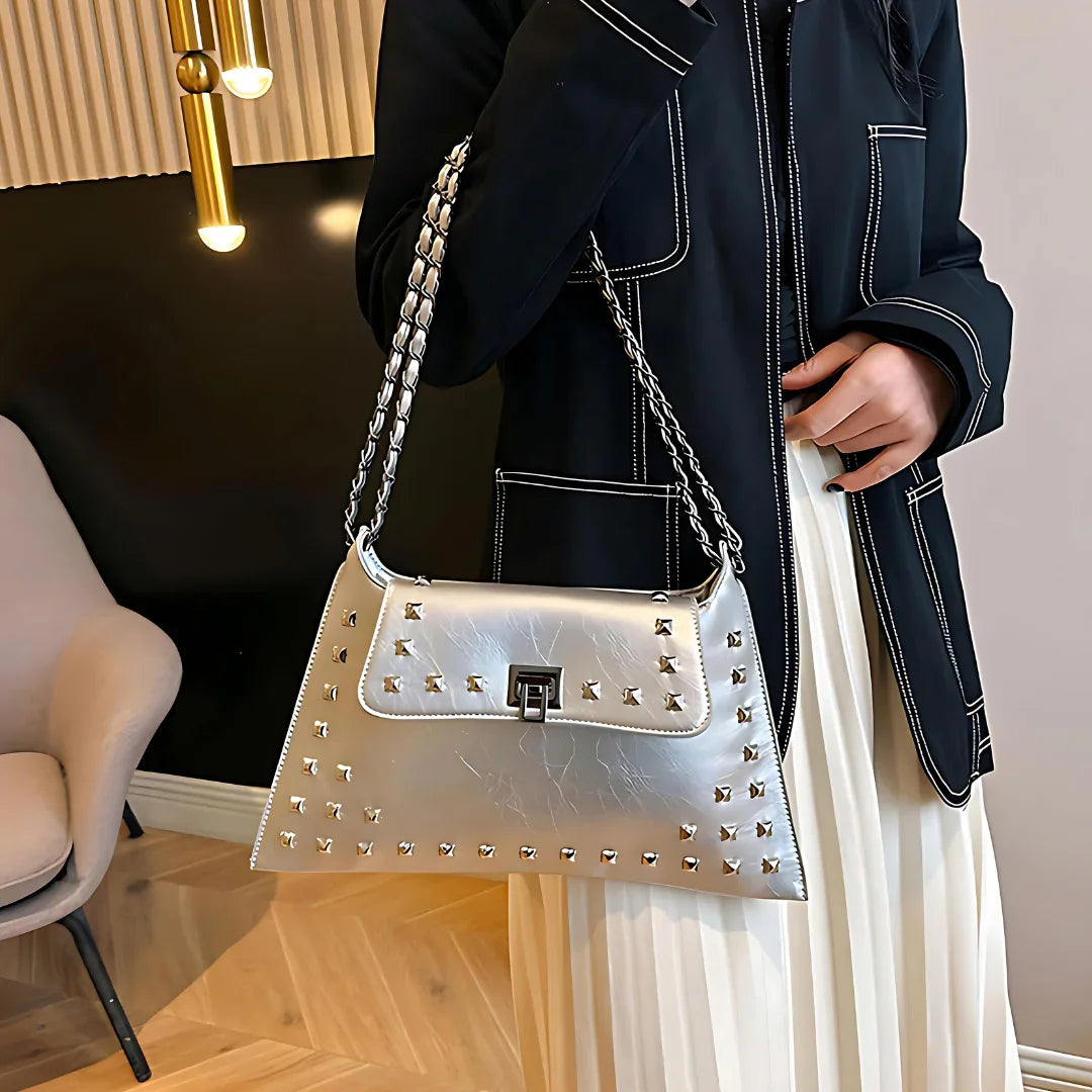 Woman holding a silver studded shoulder bag with chain strap, wearing black jacket and white skirt