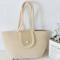 Beige woven tote bag with pearl button, double handles, on white table with flowers.