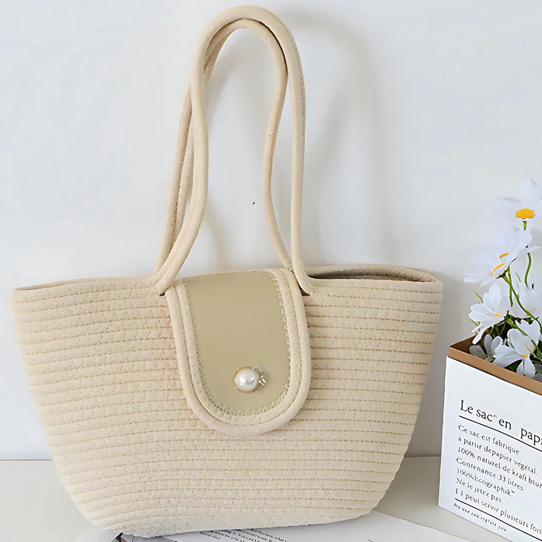 Beige woven tote bag with pearl button, double handles, on white table with flowers.