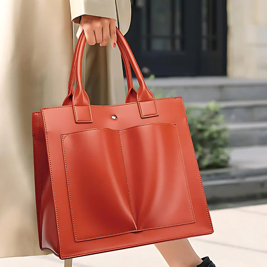 Woman holding large orange leather tote bag with front pockets, outdoor urban setting