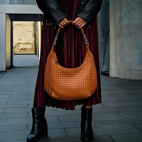 Woman holding woven brown leather hobo bag, wearing black boots and burgundy pleated skirt