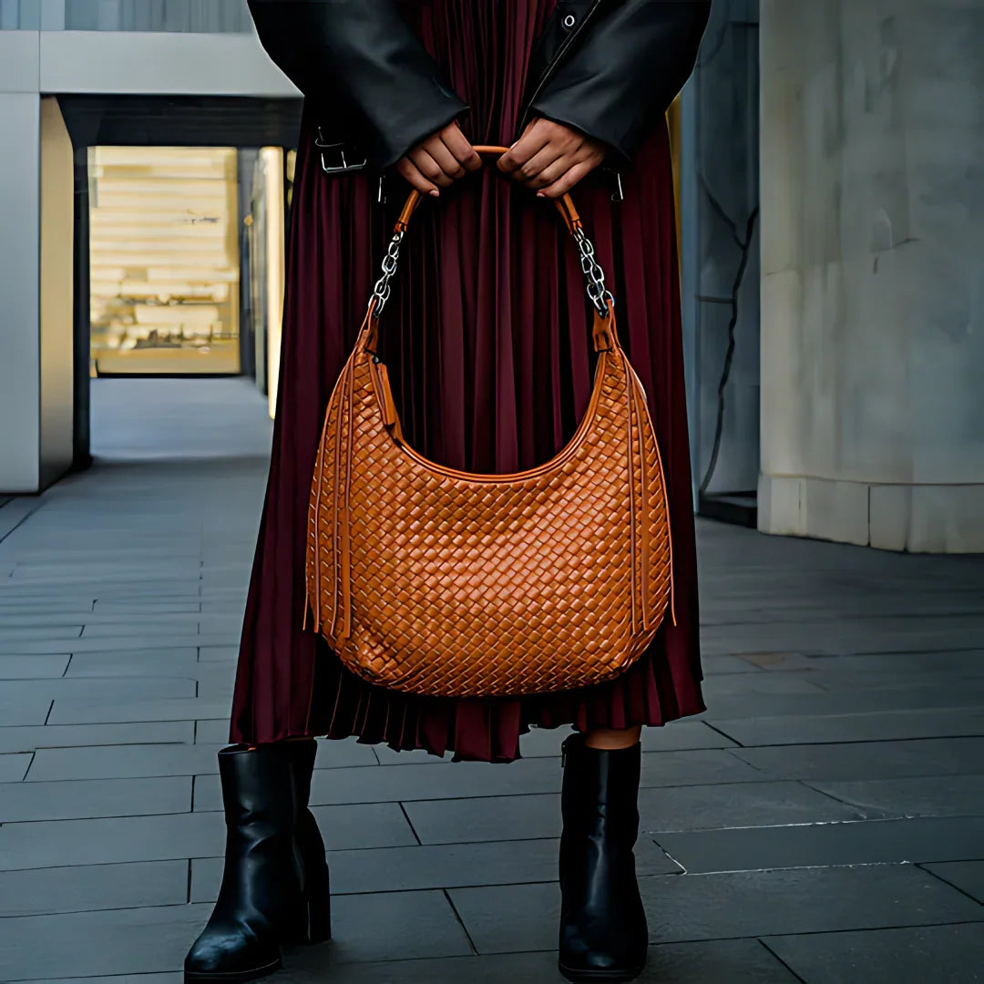 Woman holding woven brown leather hobo bag, wearing black boots and burgundy pleated skirt