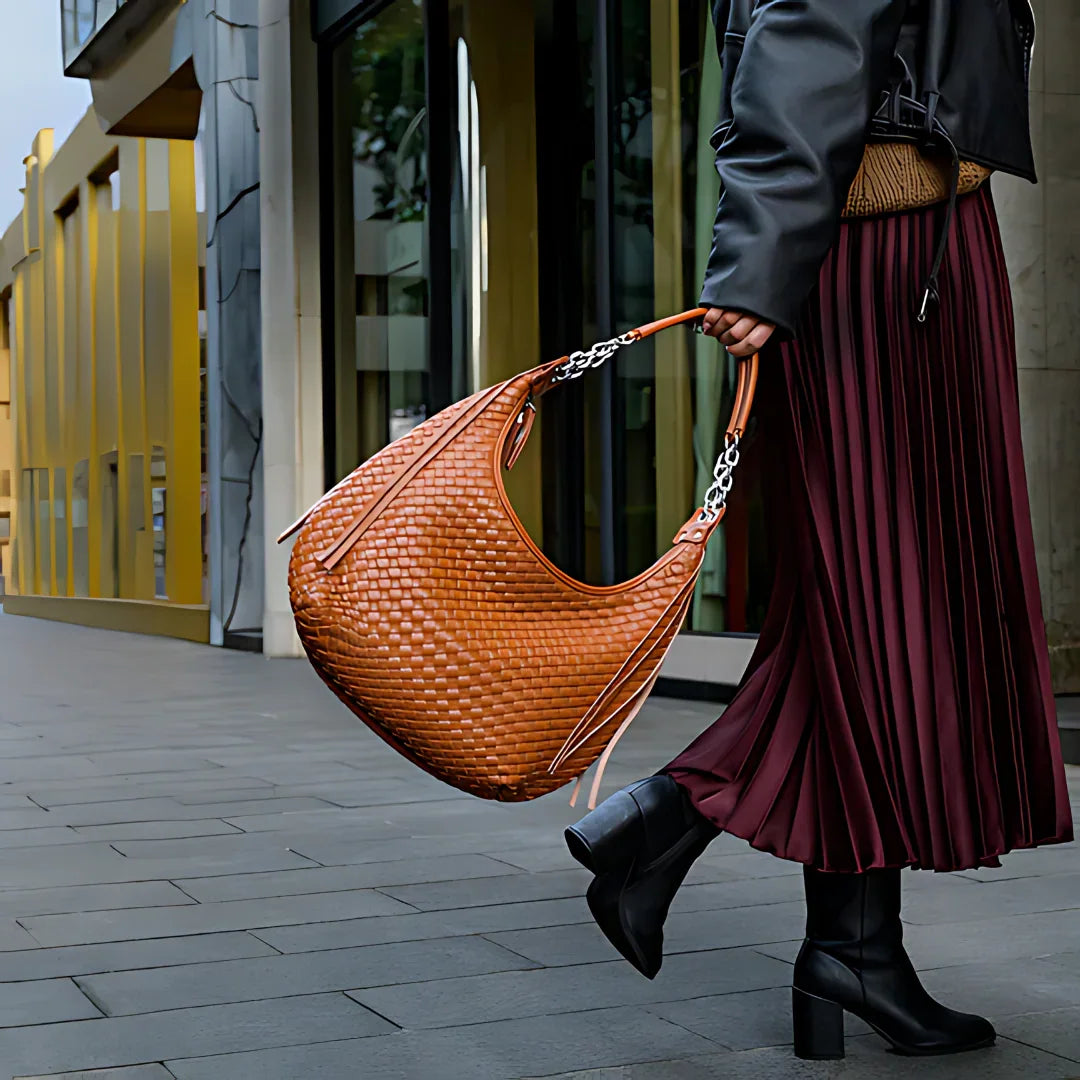 Woman with large woven brown leather hobo bag, pleated skirt, and black boots on city street