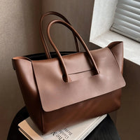 Brown leather tote bag with double handles on a table by a window, luxury handbag style