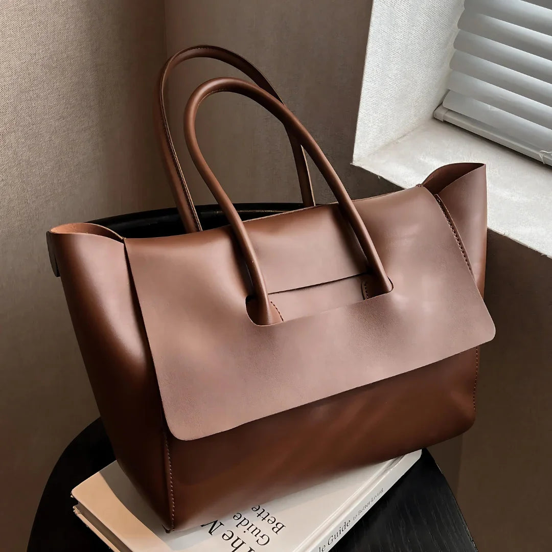 Brown leather tote bag with double handles on a table by a window, luxury handbag style