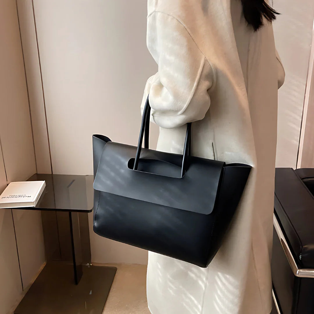 Woman in beige coat holding black minimalist leather tote bag indoors