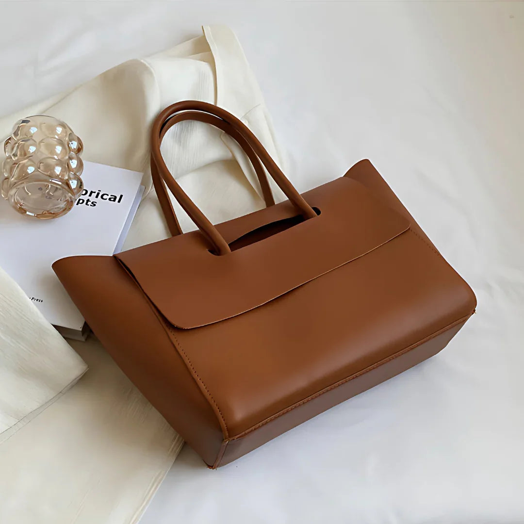 Brown leather handbag on white fabric beside a book and glass decor, minimalist style