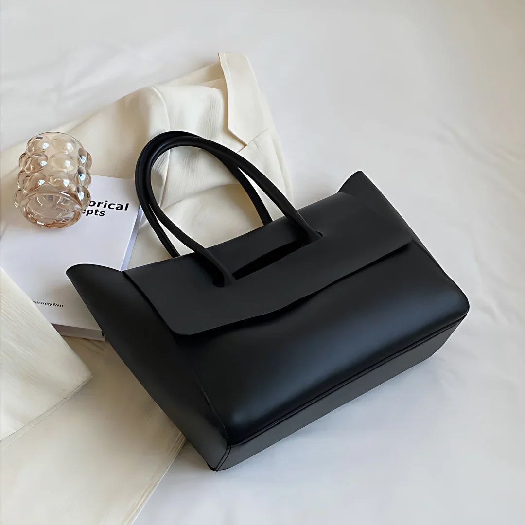 Black leather tote bag on white fabric with book and glass accessory