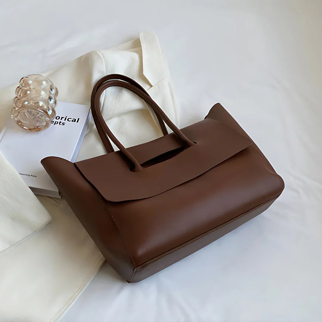 Minimalist brown leather tote bag on white fabric with a book and clear glass cup