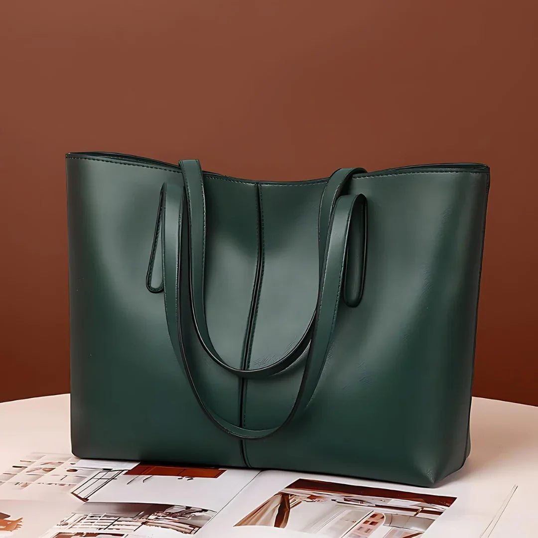 Dark green leather tote bag with double handles on a table with magazines, brown background