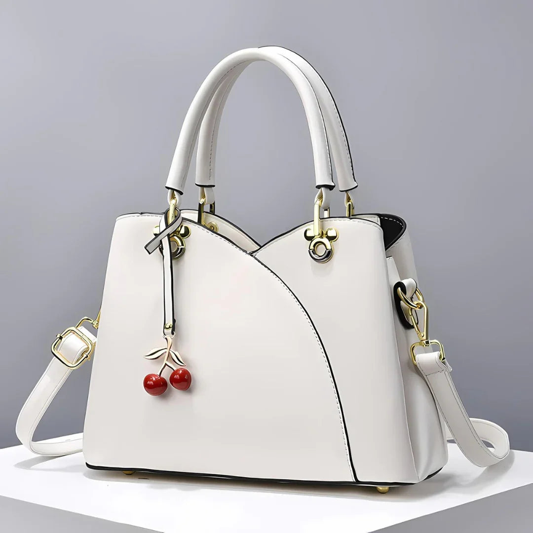 White leather handbag with gold hardware and cherry charm on neutral background