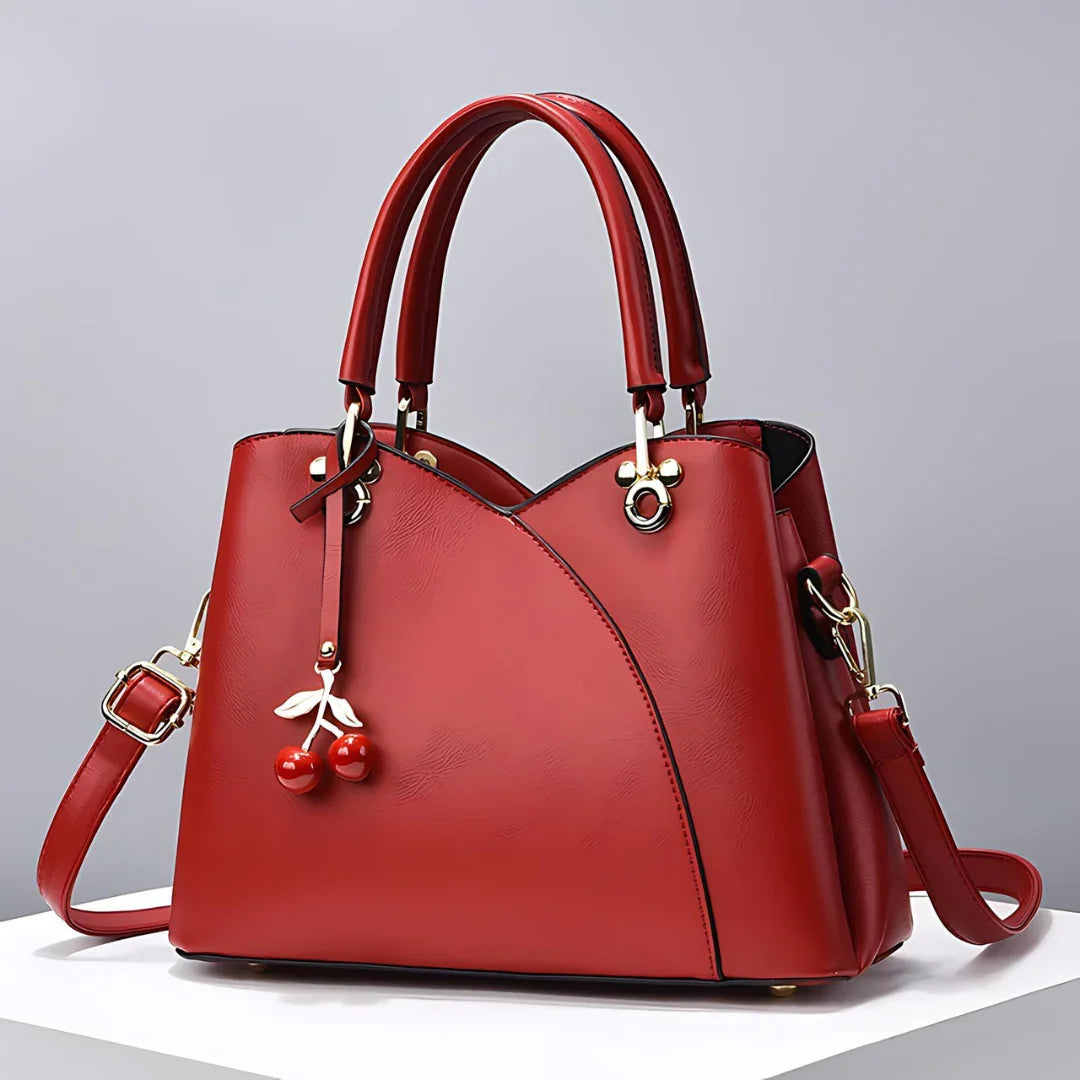Red leather handbag with cherry charm, gold hardware, and detachable strap on white stand