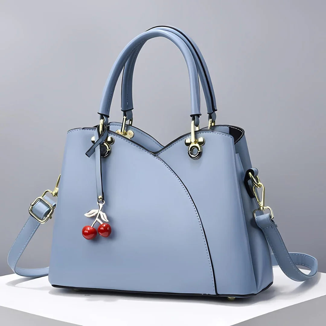 Light blue handbag with double handles, shoulder strap, and cherry charm on white display