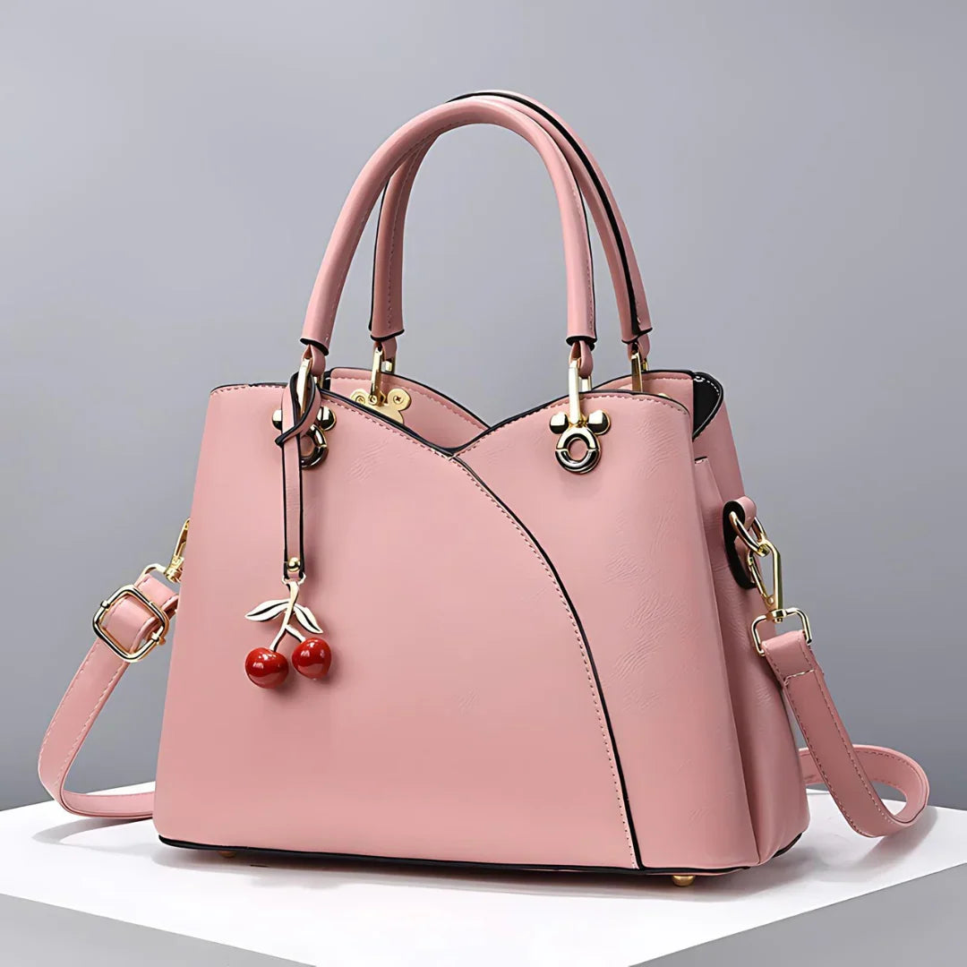 Pink leather handbag with gold hardware and cherry charm on white podium