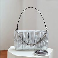 Silver pleated shoulder bag with chain strap on white table, extra strap beside it