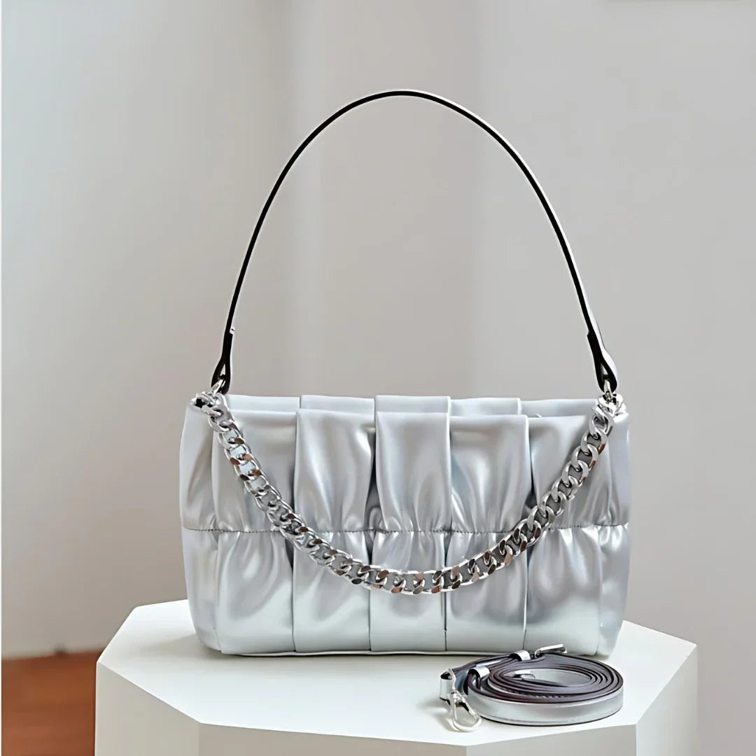 Silver pleated shoulder bag with chain strap on white table, extra strap beside it
