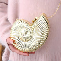 Gold seashell clutch purse with chain held by hand against soft pink clothing.