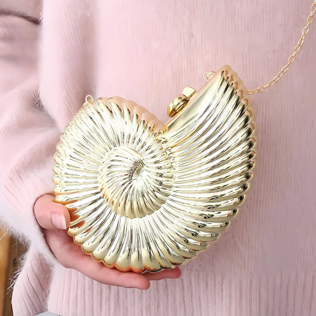 Gold seashell clutch purse with chain held by hand against soft pink clothing.