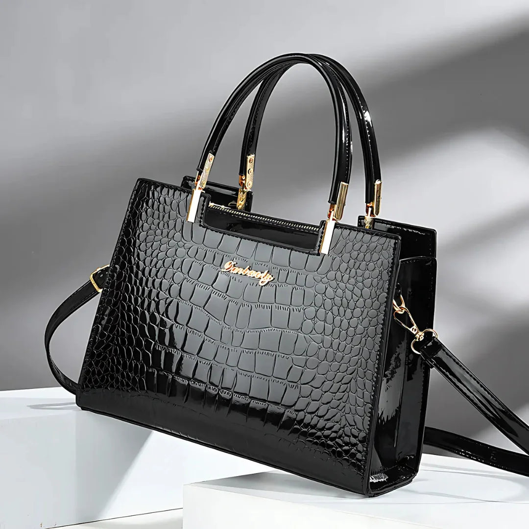 Black crocodile pattern handbag with gold hardware and dual handles on a white platform