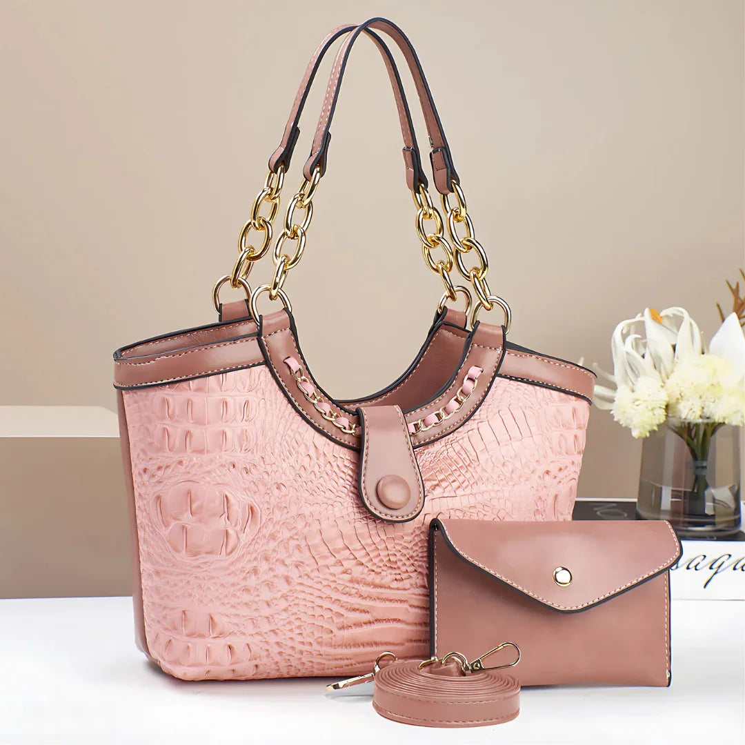 Pink crocodile-embossed handbag set with gold chain handles, wallet, and keychain on table