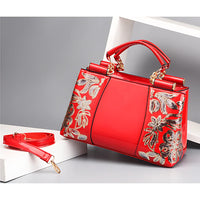 Red patent leather handbag with gold floral embroidery and detachable strap on gray background