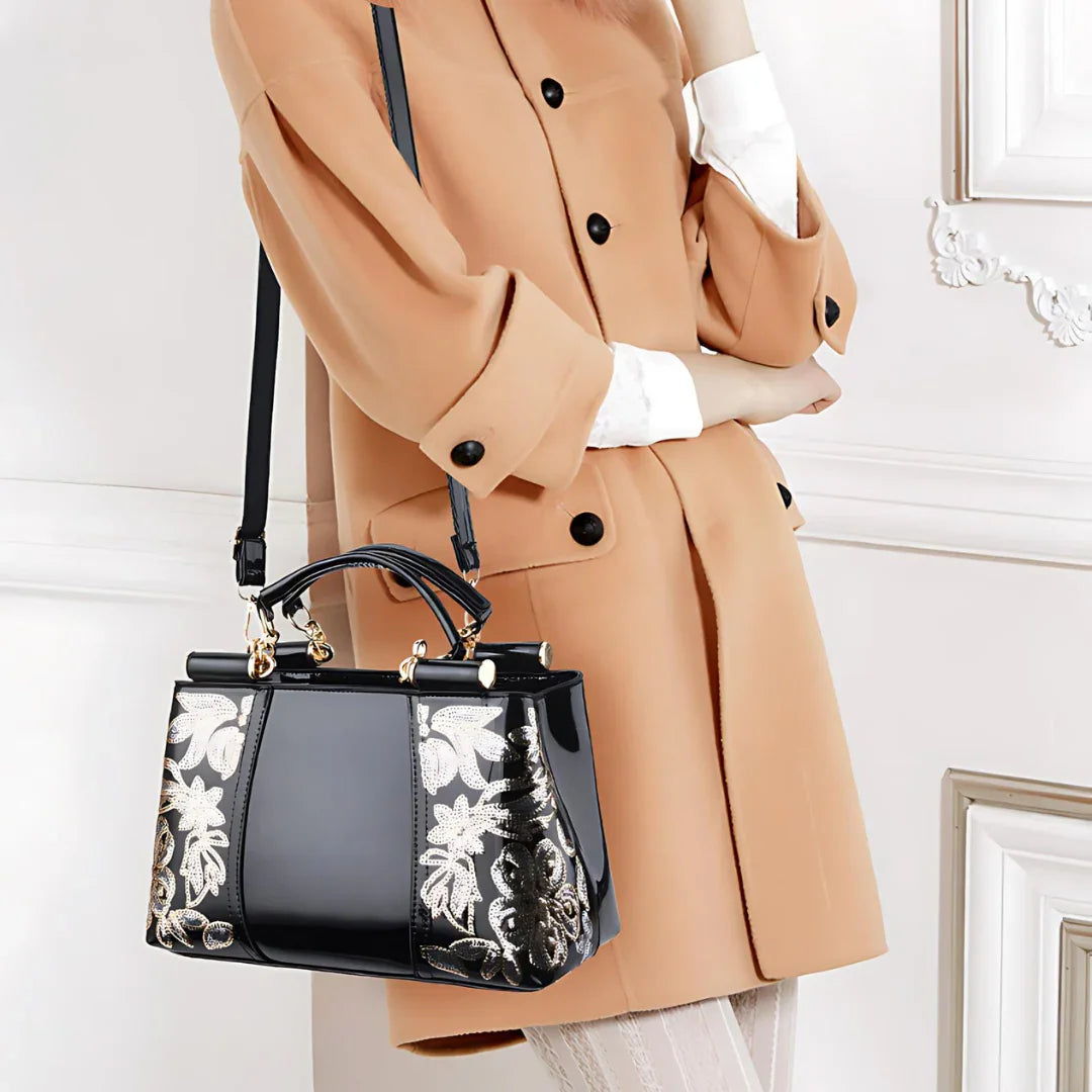 Woman in camel coat holding black patent leather handbag with gold floral embroidery