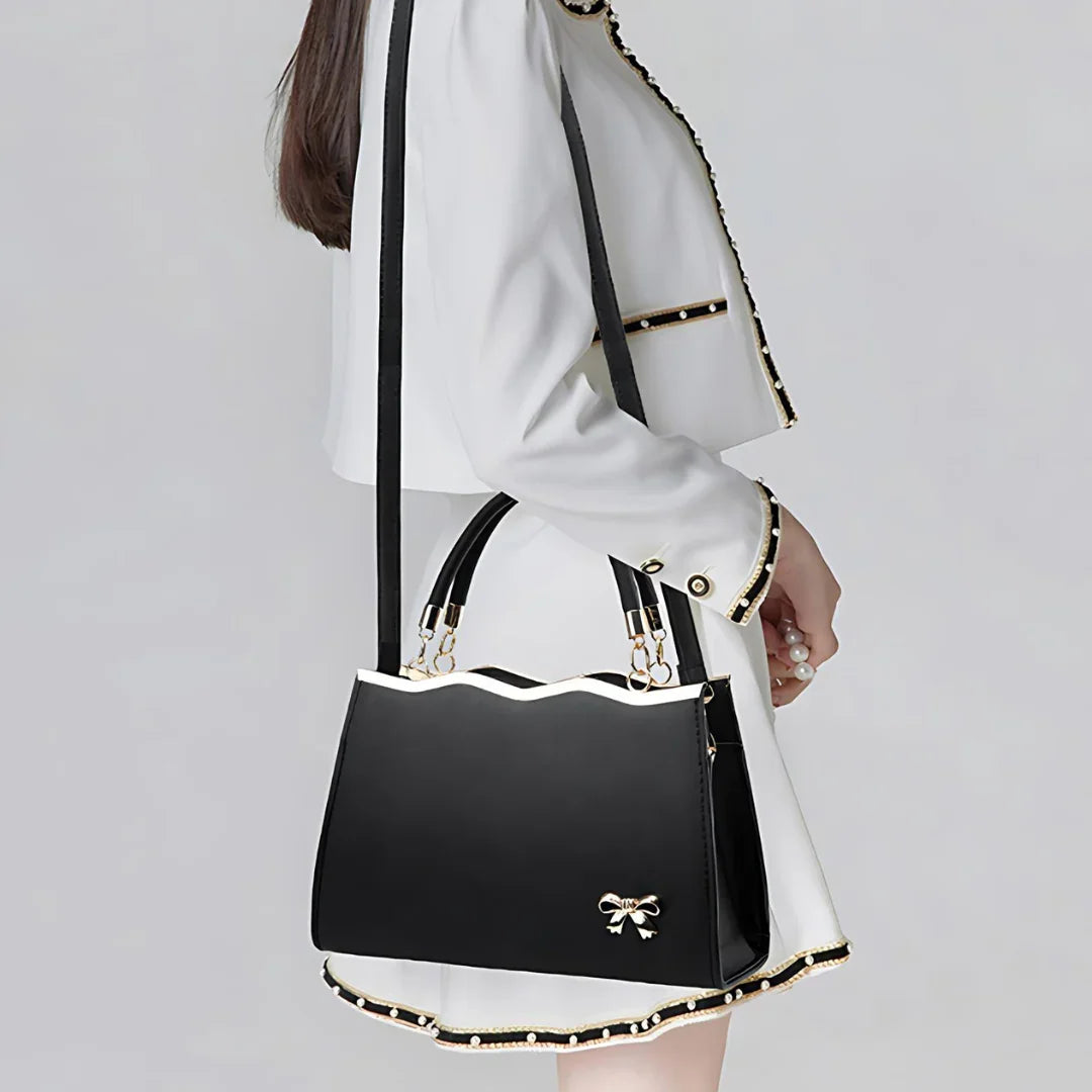 Woman in white blazer and skirt holding black handbag with gold bow charm and scalloped edge