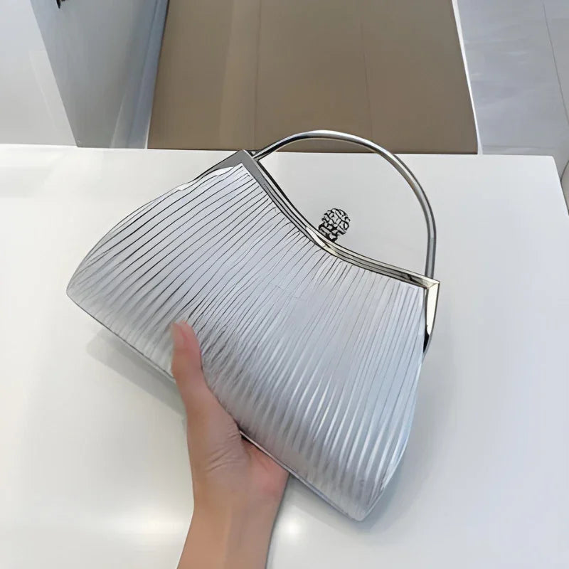 Hand holding a silver metallic pleated handbag with top handle on white table