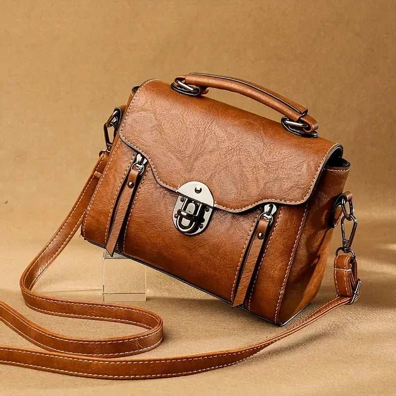 Brown leather crossbody handbag with top handle and metal buckle on tan background
