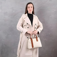 Woman in beige trench coat holding a white handbag with brown straps, grey background