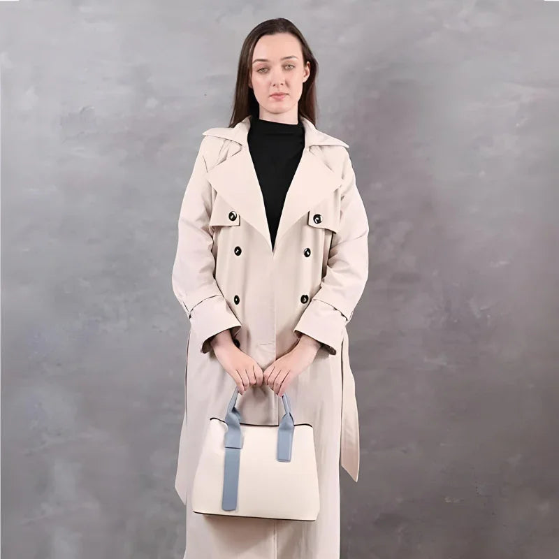 Woman in beige trench coat holding cream handbag with blue straps, gray background